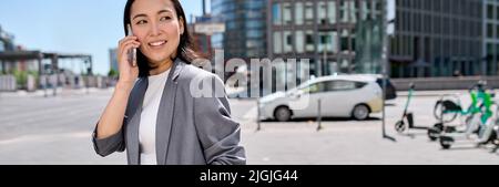 Young happy Asian business woman wears suit talking on the phone on city street. Stock Photo