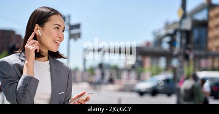 Young Asian woman talking on phone by wireless earphones on city street. Stock Photo