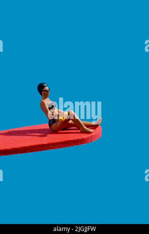 closeup of a miniature woman, wearing a black bikingi, a swimming cap and googles, sitting on a red diving board, on a blue background Stock Photo