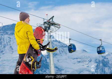 Back view of female snowboarder standing with snowboard in one hand and enjoying alpine mountain landscape - snowboarding concept Stock Photo