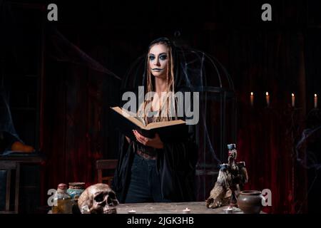 Halloween concept. Witch dressed black hood with dreadlocks standing dark room use magic book conjuring magic spell. Female necromancer wizard gothic Stock Photo