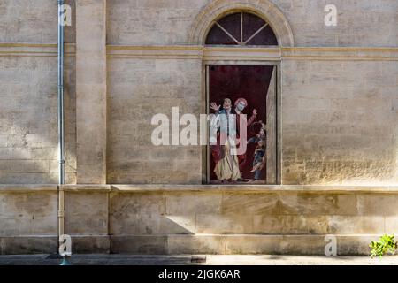 A painted window shows two masked theatrical figures in red and white-and-blue costume with two children in front of a red door, on a façade in the old town of Avignon, France. Since 1986, artist Marion Pochy and artist Dominique Durand have painted around 70 such windows across the city with motifs from past productions of the Avignon Theatre Festival, founded by Jean Vilar in 1947. Several of these windows were donated by the company JCDecaux. Stock Photo