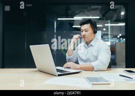 Successful asian employee boss businessman working with laptop using headset for video call, man at work in office Stock Photo