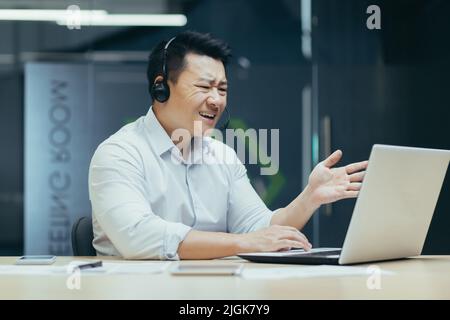 Angry and annoyed asian businessman with headset for video call, talking in online meeting, looking at laptop screen, man at work Stock Photo