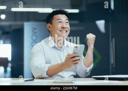 Happy and successful Asian businessman working in office, typing on phone and reading good news, celebrating triumph and victory Stock Photo