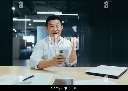 Successful asian businessman with phone looking at camera and smiling, celebrating victory, man at work in office Stock Photo
