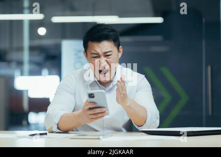 Angry businessman in office yelling on the phone, Asian man frustrated with received email Stock Photo