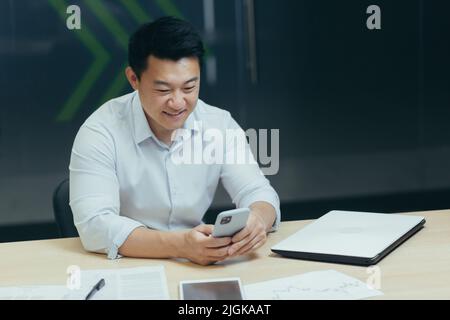Asian man using phone in modern office, typing message and reading news while sitting at desk, businessman at work Stock Photo