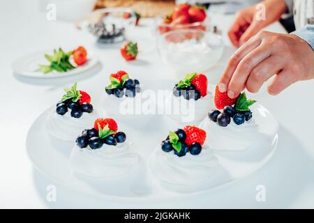 Cooking, making homemade meringue. Female hands decorating pavlova cakes with fresh berries on the kitchen table with ingredients. Recipes for delicio Stock Photo