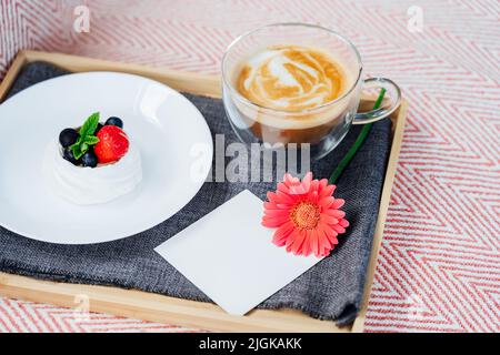 Cup of coffee, Pavlova dessert cake, gerbera flower and blank postcard with copy space on the tray with gray napkin. Breakfast for loved one. Romantic Stock Photo
