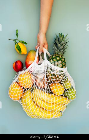 Top view female hand holding string bag, mesh bag, grocery bag with organic fresh exotic fruits on light green background. Zero waste, eco-friendly sh Stock Photo
