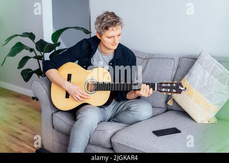 Man taking online guitar lessons via a mobile application on his phone at home sitting on the gray couch, sofa in the living room. He watching acousti Stock Photo