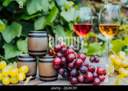 Winery concept. Miniature wine barrels, glasses with white and red wine and grape berries on the wooden table on background of vineyards. Wine tasting Stock Photo