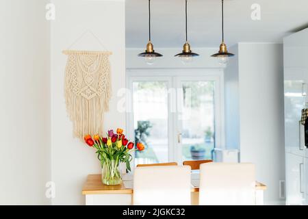 Stylish and modern boho, scandi interior of open space white kitchen with tulip flowers in vase on the wooden kitchen counter table, beige macrame and Stock Photo