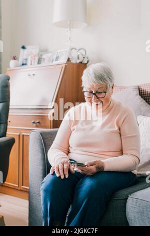 Portrait of happy laughing senior woman using phone while sitting onon the sofa at home. Mature older lady is excited to receive good news via smartph Stock Photo