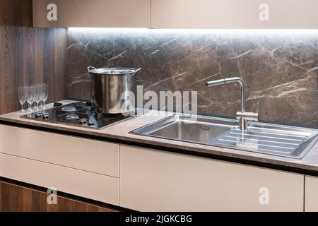 Closeup gas stove with metal saucepan and washbasin with tap located on counters near set of wineglasses in modern kitchen Stock Photo