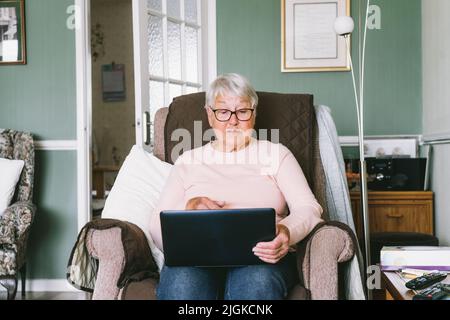 Senior grandmother sitting on armchair, looking at laptop screen. Grey-haired mature woman reading news, chatting with family, watching video, online Stock Photo