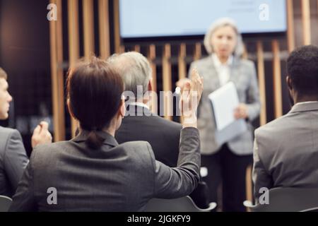 Rear view of businesswoman in jacket sitting in auditorium and raising hand while having question to conference speaker Stock Photo