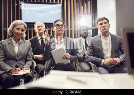 Serious young businesswoman in glasses sitting among conference participants and raising hand while having question to marketer after presentation Stock Photo