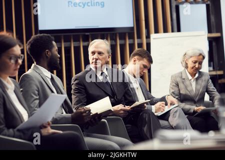 Group of multi-ethnic entrepreneurs in suits sitting in row at conference on recycling business and summarizing information Stock Photo