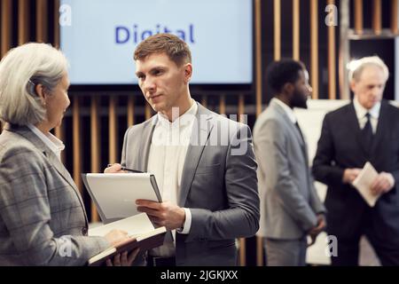 Serious handsome young journalist in suit making notes in clipboard while interviewing business forum participant during break Stock Photo