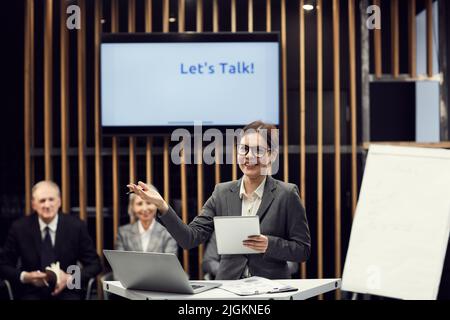 Portrait of positive excited young business lady in eyeglasses standing at speech table and producing arguments while giving speech at meeting Stock Photo