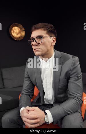 Serious young businessman in suit and glasses sitting on comfortable chair and talking to important person at interview Stock Photo