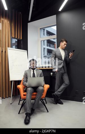 Young successful managers working with social medias: smiling black businessman sitting in orange armchair and typing on laptop while his colleague sc Stock Photo