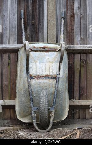Old builders wheelbarrow standing on end against old worn wooden fence Stock Photo