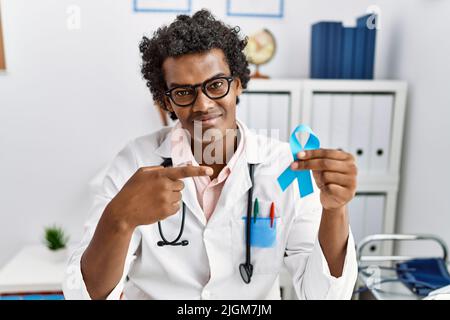 African doctor man holding blue ribbon smiling happy pointing with hand and finger Stock Photo
