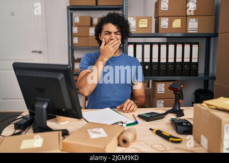 Hispanic man with curly hair working at small business ecommerce bored yawning tired covering mouth with hand. restless and sleepiness. Stock Photo