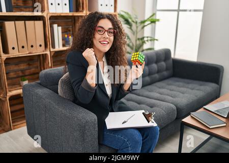 Young hispanic woman playing colorful puzzle cube intelligence game screaming proud, celebrating victory and success very excited with raised arms Stock Photo