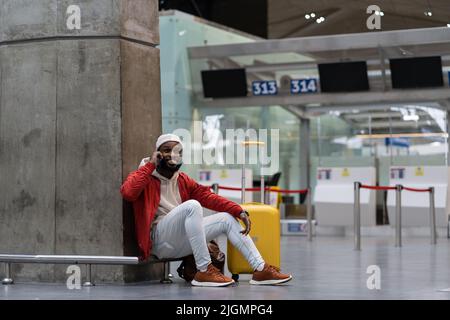 Handsome African man in red jacket calling on smartphone in airport while waiting flight in lounge. Stock Photo