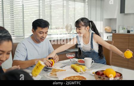 Happy asian family having breakfast at the kitchen table, daughter serving healthy fruit with father. Stock Photo