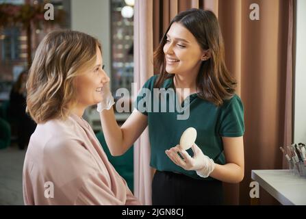 Professional makeup artist doing makeup for woman in beauty salon. Smiling woman beauty specialist in sterile gloves applying powder on client face with cosmetic brush. Stock Photo