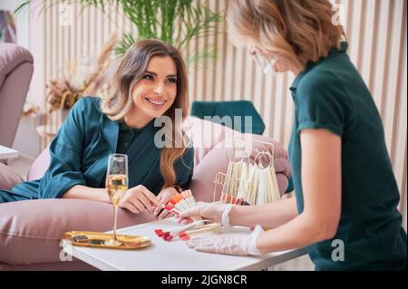 Cheerful woman pointing at red color nail sample and smiling while choosing manicure with manicurist. Manicure master in sterile gloves holding nail polish color palette. Stock Photo