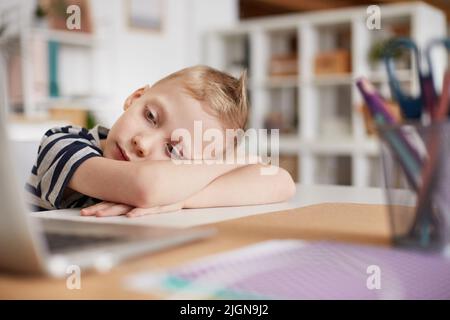 Serious tired boy with blond hair sitting at table and leaning head on crossed arms while watching informative documentary on laptop Stock Photo