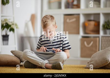Serious concentrated blond-haired boy in stripped tshirt sitting with crossed legs on carpet and using tablet while surfing internet Stock Photo