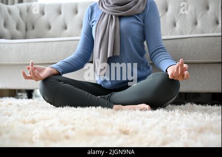 Calm and peaceful Muslim woman wearing hijab practicing yoga, relaxes meditating in her home living room. cropped image Stock Photo