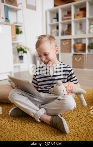 Happy excited cute boy with toy bear sitting on floor and laughing while using tablet at home Stock Photo