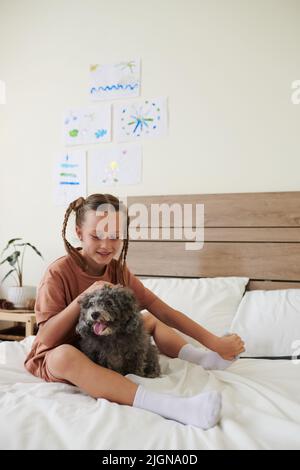 Smiling girl sitting on bed with cute curly dog and petting it on head Stock Photo