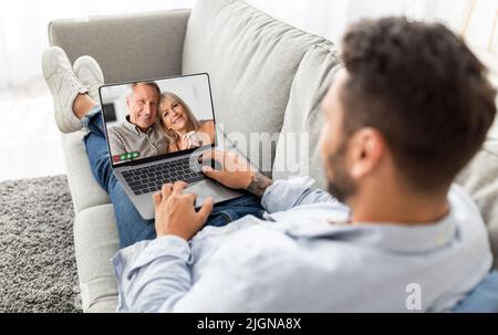 Man Making Video Call On Laptop With Senior Parents While Relaxing On Couch Stock Photo