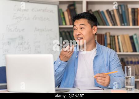 Asian Male Tutor Using Voice Search Application Sitting At Workplace Stock Photo