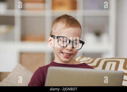 Smiling cute nerd boy in eyeglasses sitting on sofa and using laptop while searching for information Stock Photo