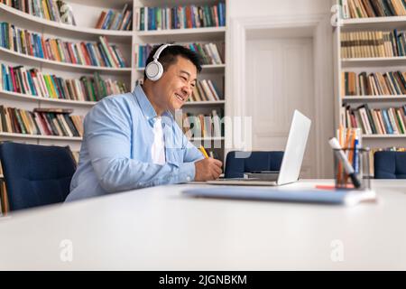 Happy Asian Man Using Laptop And Taking Notes In Library Stock Photo