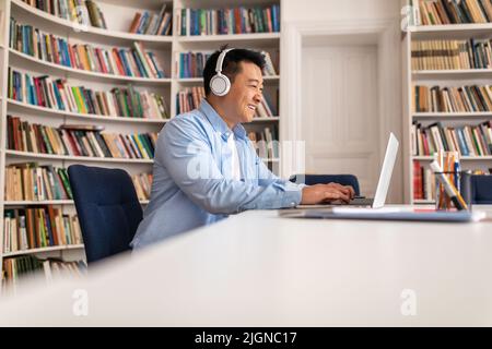 Korean Man Wearing Headphones Using Laptop Typing Sitting In Library Stock Photo