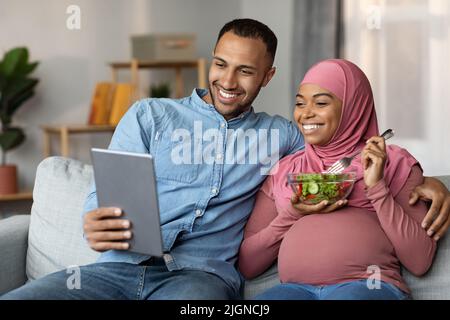Pregnant Couples Leisure. Black Islamic Spouses Relaxing With Digital Tablet On Couch Stock Photo