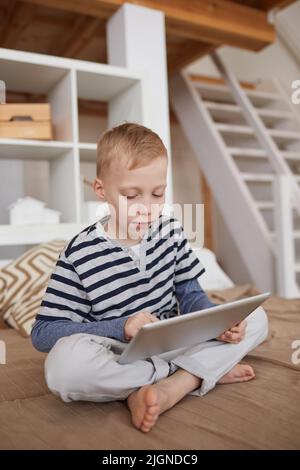 Serious blond-haired boy in casual outfit sitting with crossed legs on bed and using portable device Stock Photo