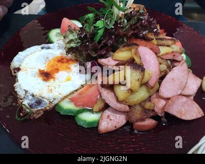 fried cut potatoes with fried cut sausages with sunny side up eggs garnish with fresh leaf salat with tomatoes and cucumber Stock Photo