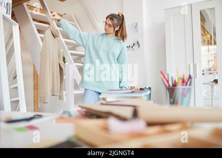 Serious attractive young female tailor in casual outfit standing in own studio and looking at finished sweater Stock Photo